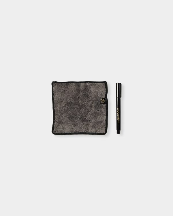 WoodUpp grey microfiber polishing cloth and a black WoodUpp touch-up marker shown on a white background. The square, plush dark-grey cloth has black bound edges and a small WoodUpp tag; the slim black marker pen features gold WoodUpp branding and a removable cap.