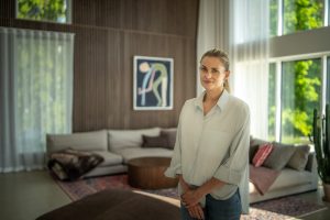 A woman stands in a bright living room featuring WoodUpp vertical wooden slat wall panels behind a neutral sofa and a WoodUpp round fluted wooden coffee table. Natural light, large windows, rugs and cushions complete the modern, warm interior shown in the photograph.