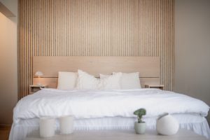 Minimal bedroom featuring WoodUpp slatted oak wall panels behind a matching WoodUpp oak headboard and two compact WoodUpp bedside tables. Crisp white bedding and textured pillows sit on the bed, with a small lamp, candles and a potted succulent on the bedside and foreground surfaces.
