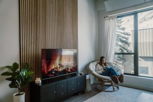 Modern living room with a floor-to-ceiling WoodUpp vertical wood slat wall panel in natural oak behind a TV showing a fireplace. A black media console, potted plant, and a woman reading in a cushioned papasan chair by a large window complete the cozy scene.